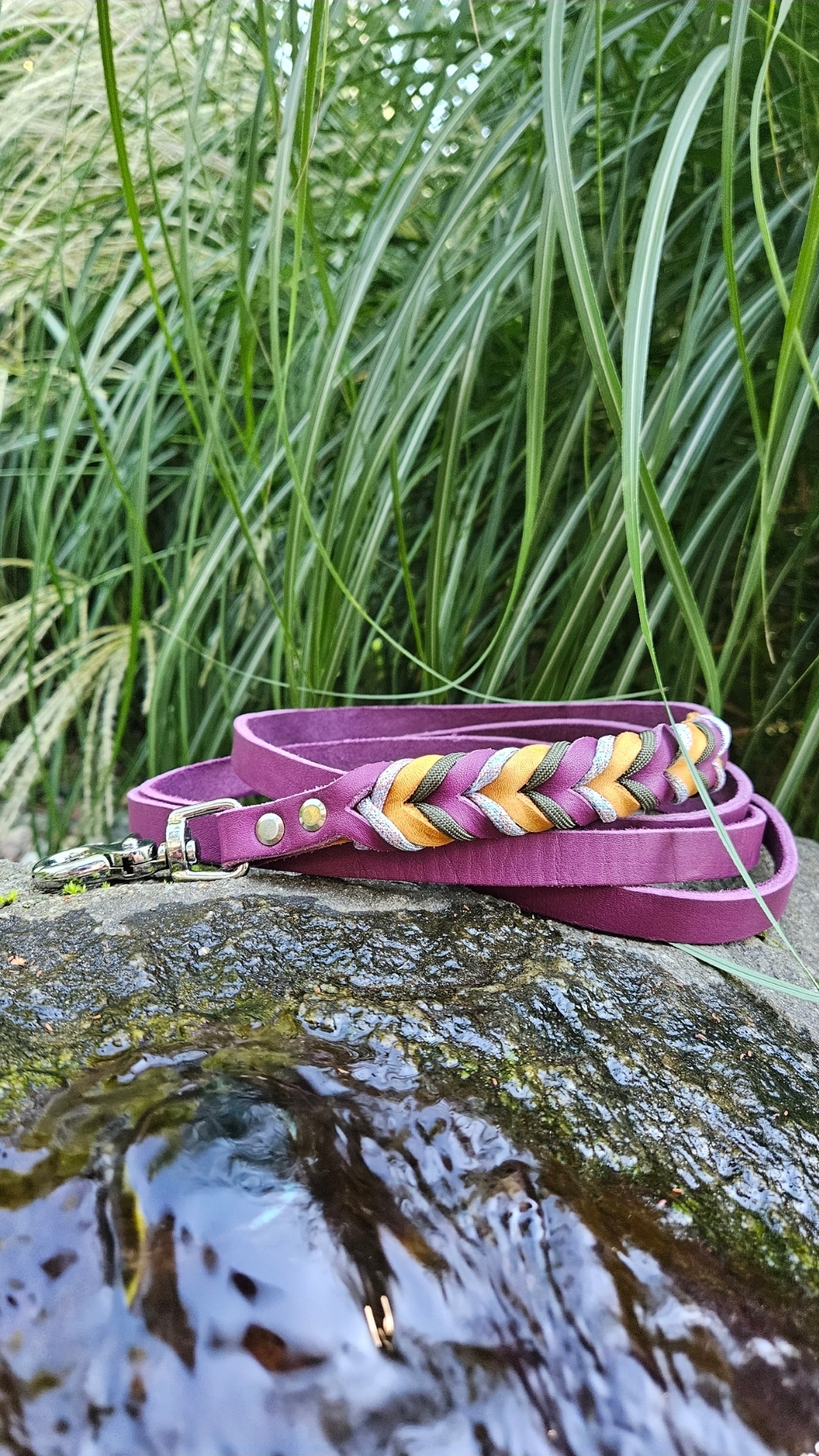 Smycz Woven Line — skórzany pasek w kolorze głębokiego fioletu z plecionką w jodełkę z żółtego, srebrnego i oliwkowego sznurka, zwinięty na mokrym kamieniu