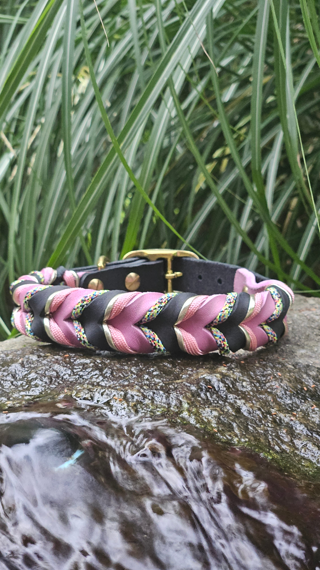 Obroża Woven Line — splot dwóch kolorów skóry z dwoma wzorami sznurka paracord i ozdobnym rzemykiem, leżący na mokrym kamieniu wśród traw